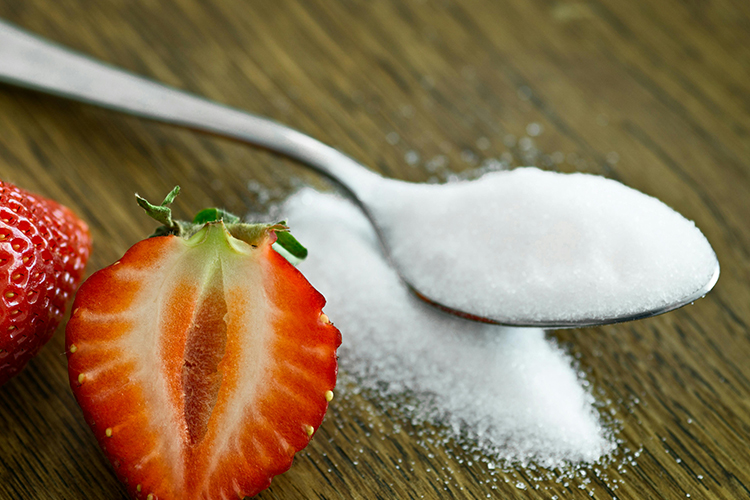 Closeup photo of a sugar-filled tea spoon on a wooden table next to 2 strawberries on the left, one of which is cut in half (sagittal cut), giving us a nice view of the inside structure.  The spoon is overloaded with sugar to such an extent that there is almost just as much that has spilled on the table.
